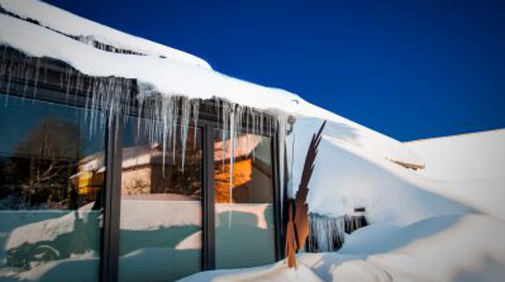 Ein moderner Wintergarten mit großen Glasflächen, bedeckt von Schnee und Eiszapfen, der eine winterliche Atmosphäre vermittelt. Im Hintergrund spiegeln sich Bäume und Gebäude.