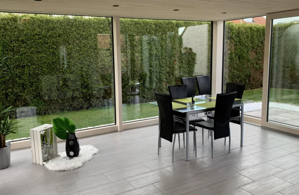 Geräumiger und lichtdurchfluteter Innenraum eines Wintergartens mit klaren Sichtlinien zum grünen Außenbereich, minimalistisch eingerichtet mit einem modernen Esstisch und Dekorationen.