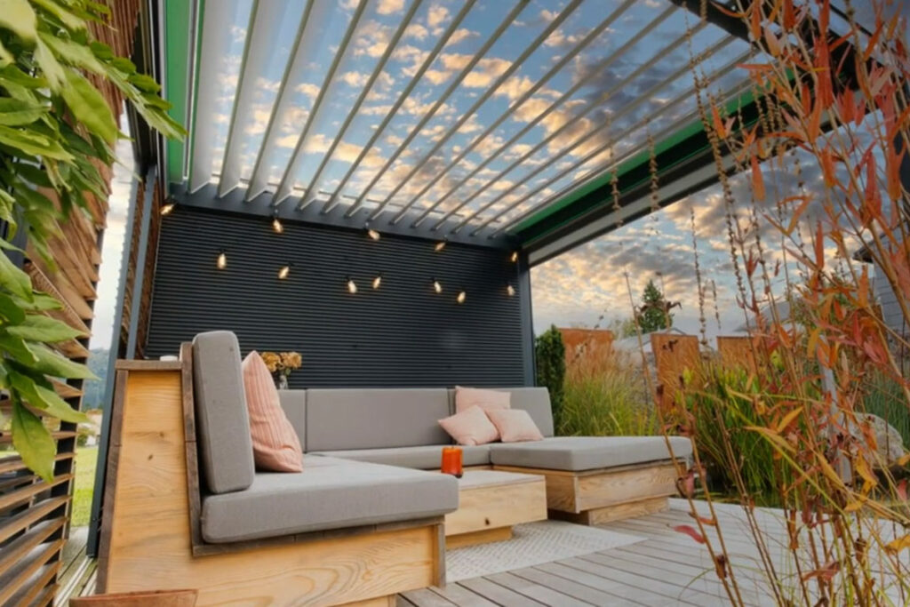 Stilvoll eingerichtete Terrasse mit einer zeitgenössischen Terrassenüberdachung aus Lamellen, die eine gemütliche Lounge-Ecke mit einem Sofa schützt. Direkt angrenzend befindet sich ein natürlicher Schwimmteich, der mit einer vielfältigen Pflanzenwelt und sanftem Grün eine beruhigende Atmosphäre schafft.