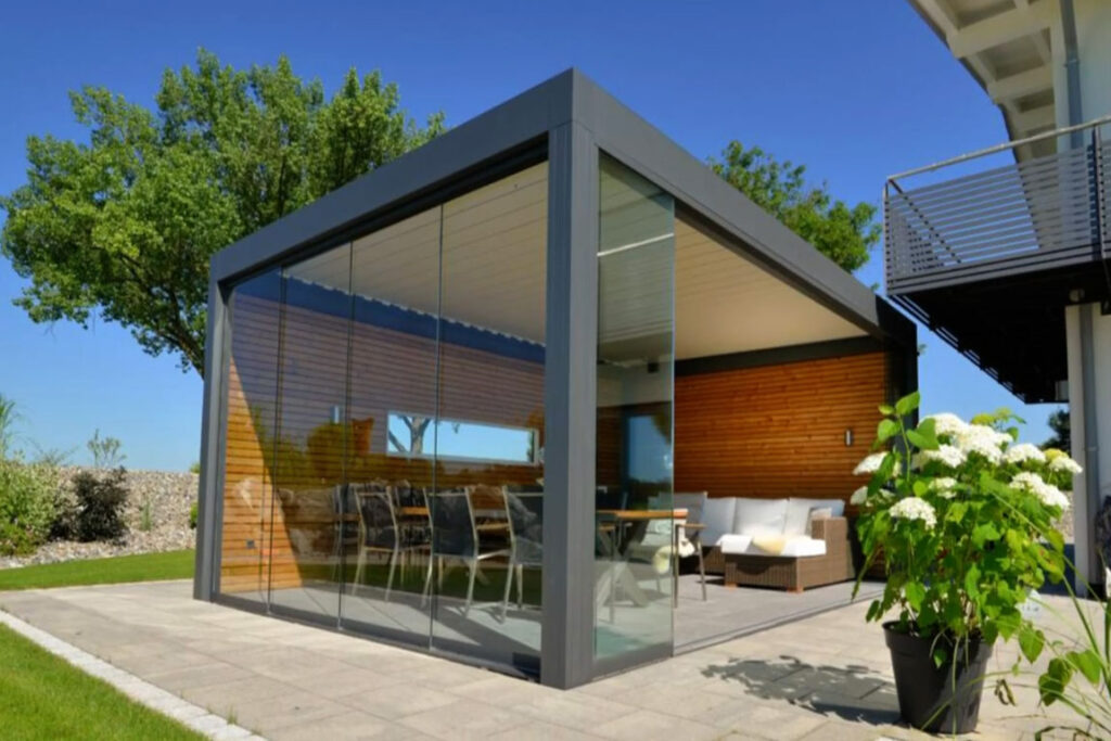 Moderne Gartengestaltung mit einem freistehenden Lamellendach über einer Terrasse, die mit einem gemütlichen Lounge-Set ausgestattet ist. Die Terrasse ist von einem gepflegten Rasen und einer vielfältigen Gartenbepflanzung umgeben.