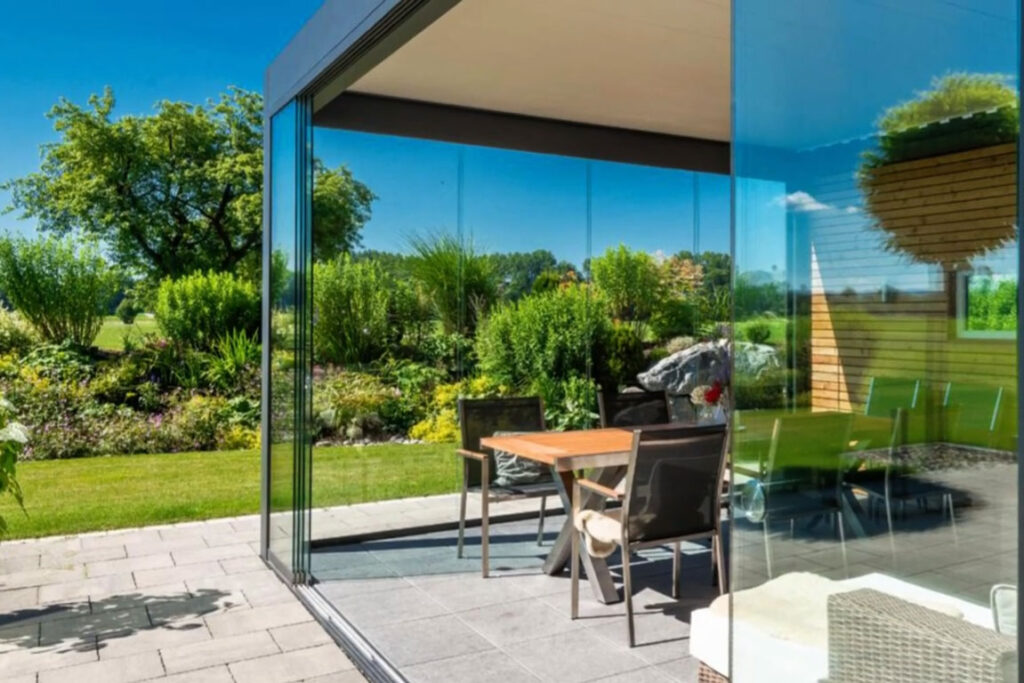 Ein einladender Gartenbereich mit einem freistehenden Lamellendach, darunter eine stilvolle Terrasse mit Outdoor-Möbeln zum Entspannen. Die Terrasse öffnet sich zu einem großzügigen, grünen Rasen und ist umrahmt von einer abwechslungsreichen Gartenanlage.