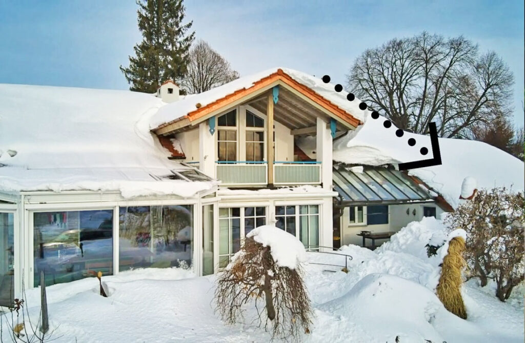 Ein verschneites Haus mit Wintergarten und einer markierten Dachkante, die auf die Gefahr einer Dachlawine hinweist. Schneebedeckte Bäume und ein klarer Himmel im Hintergrund.