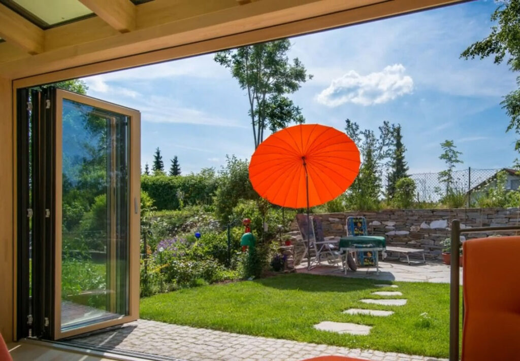 Geöffnete Glas-Falttür mit Blick auf einen sonnigen Garten mit Steinweg, Gartenmöbeln und einem großen orangefarbenen Sonnenschirm.