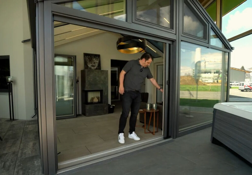 Matthias Brack steht in einem modernen Wintergarten und schiebt eine große Glas-Schiebetür zur Seite, um den Raum zur Terrasse zu öffnen.