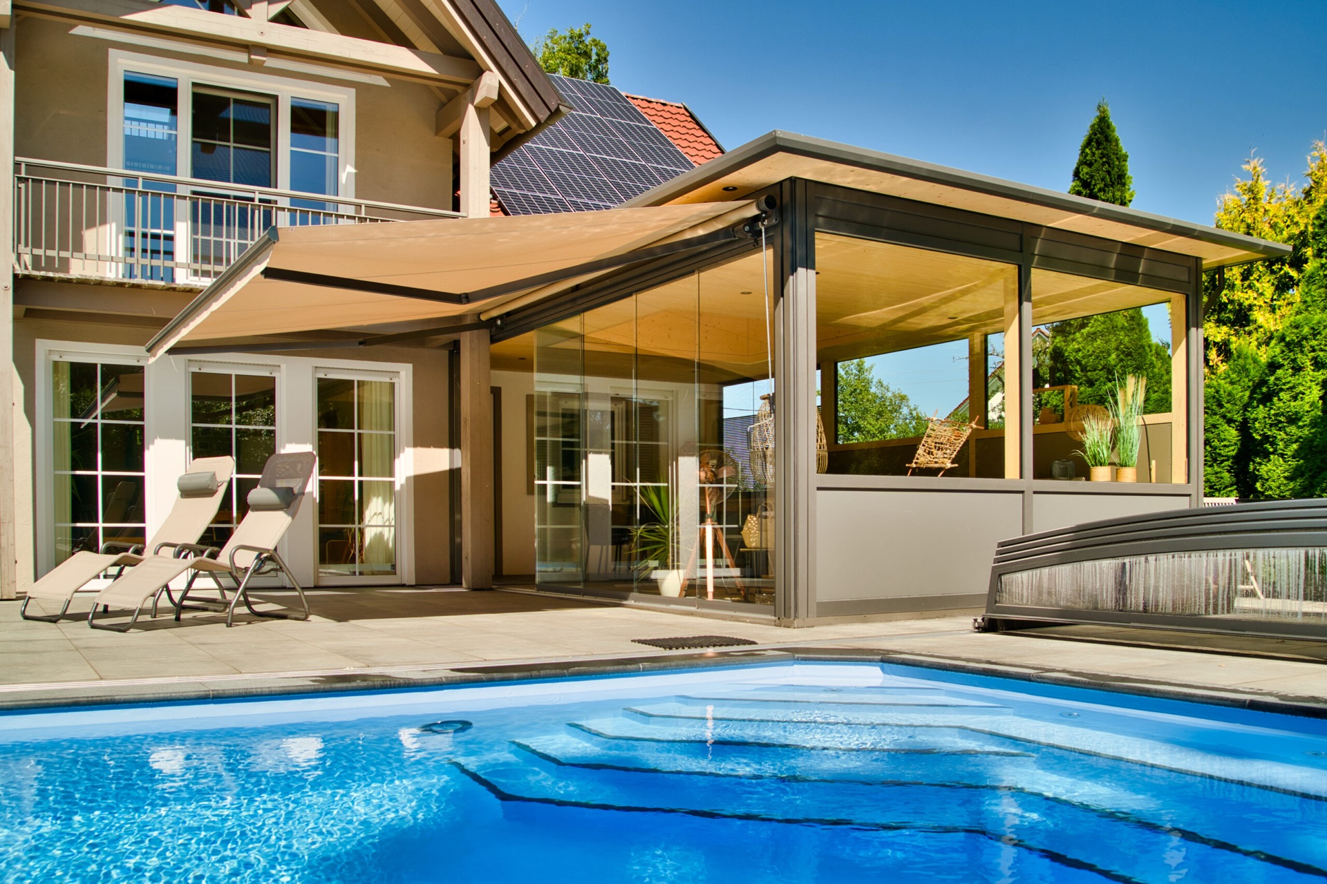 Überdachte Terrasse mit flacher, beiger Markise direkt an einem Wintergarten neben dem Swimmingpool.
